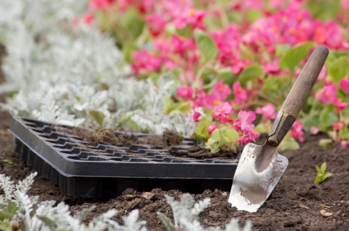 A gardening scene in a planted outdoor area features a black seed tray resting on dark, moist soil, with a small garden trowel inserted into the ground nearby. The background displays a vibrant mix of pink and white flowering plants and green foliage, suggesting a well-maintained garden space typical of outdoor gardening services in Earls Court. The soil appears freshly turned, indicating recent planting or soil preparation, with a natural, slightly textured surface. The seed tray is partially filled with soil or compost, ready for sowing seeds, and the garden trowel has a wooden handle and a metal blade, showing signs of use. The overall setting is outdoors under natural daylight, with a lush, colorful garden environment that highlights the work involved in garden maintenance and planting tasks in a typical London backyard or front garden, aligning with gardening services offered by Gardening Services Earls Court. The arrangement signifies ongoing or upcoming planting activities suitable for professional gardening care in the Earls Court area.