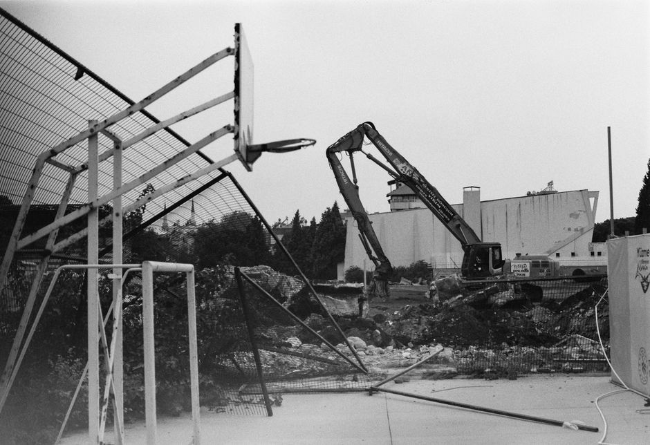 A black-and-white image depicting a construction site where landscaping work is in progress, featuring an excavator with a hydraulic arm digging into the ground near a fenced-off area. In the foregrou